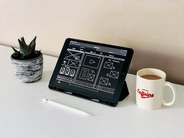 Tablet displaying wireframe designs for a website redesign on a desk with a potted plant, stylus, and coffee mug.