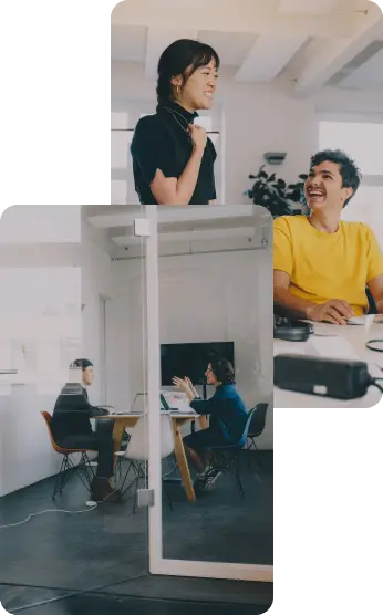 People are engaged in a conversation in a bright, modern office. One person stands, while others sit around a table with laptops.